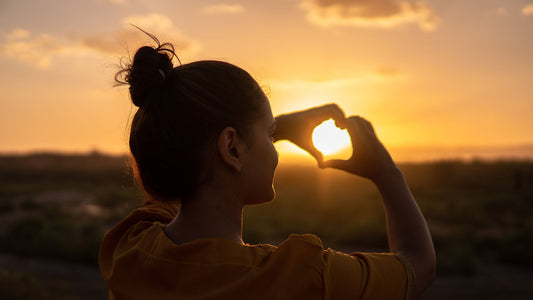 Woman making a heart sign in front of a sunset